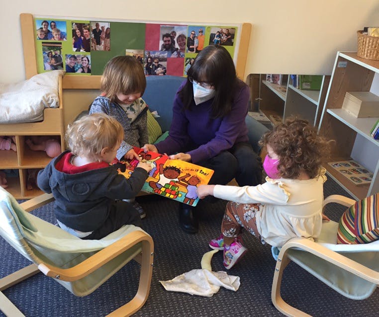 Person wearing mask reading book to three children in a classroom