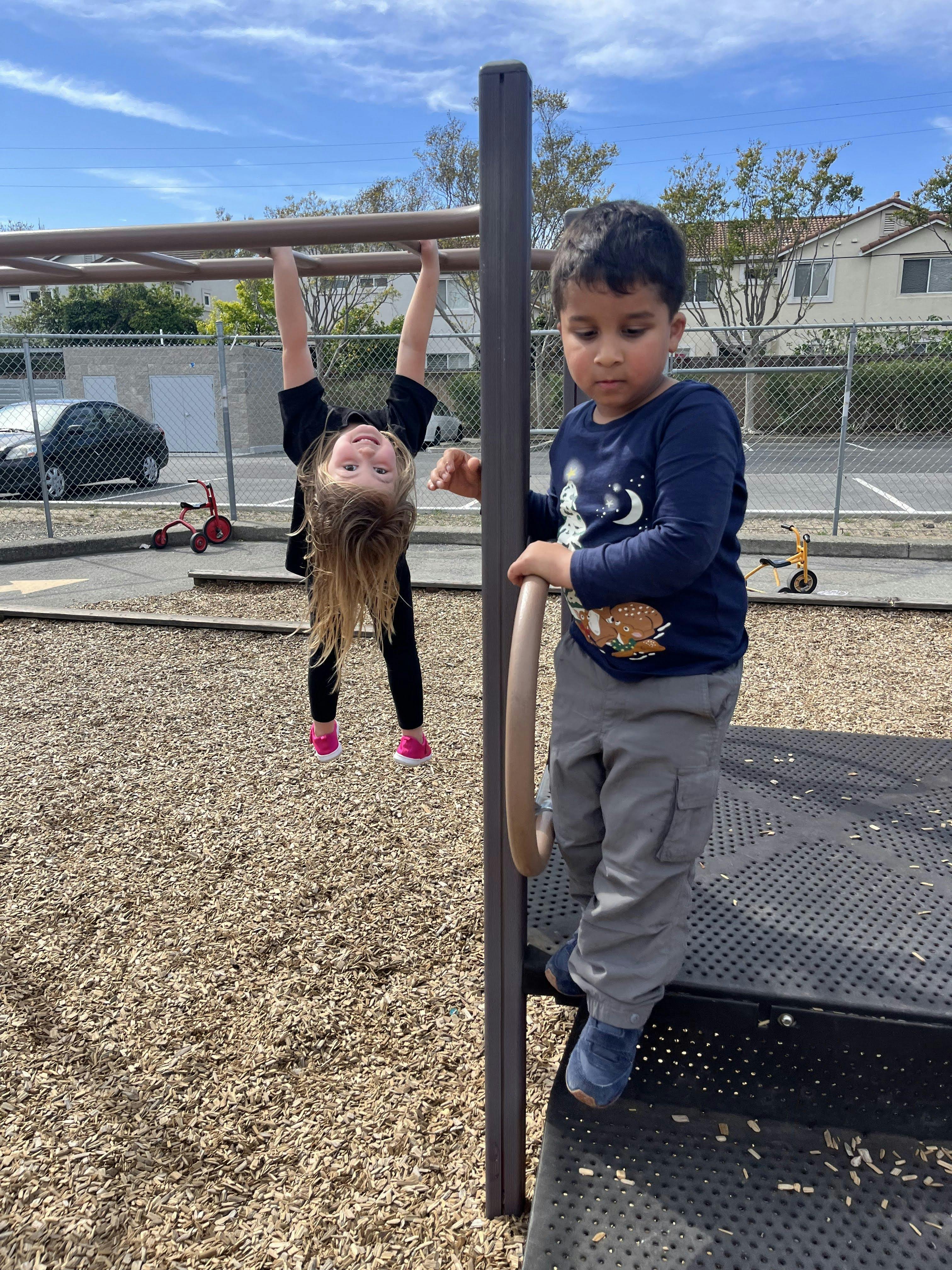 Child hanging upside down from monkey bars, another child on playground equipment