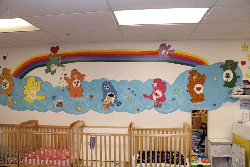Nursery room with cribs and a wall mural of cartoon bears on clouds and a rainbow