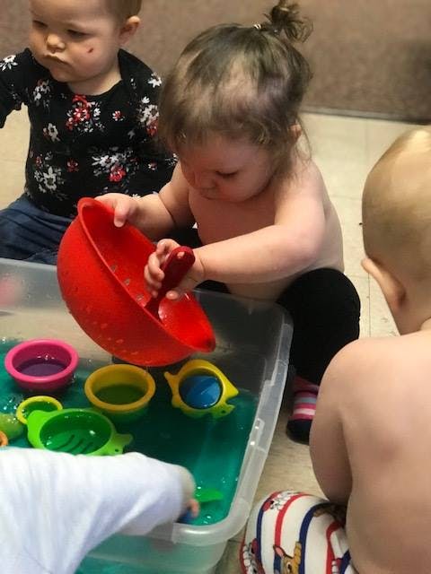 Child playing with a red bowl and spoon in a bin of water with other children