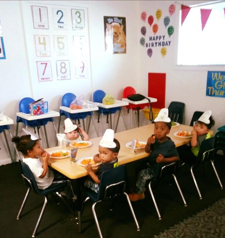 Children wearing paper hats eating at a long table in a classroom