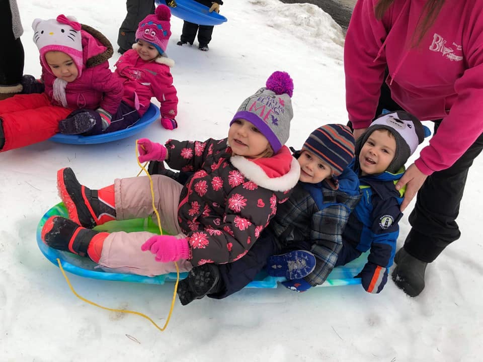 Three children on a sled in the snow with an adult standing nearby