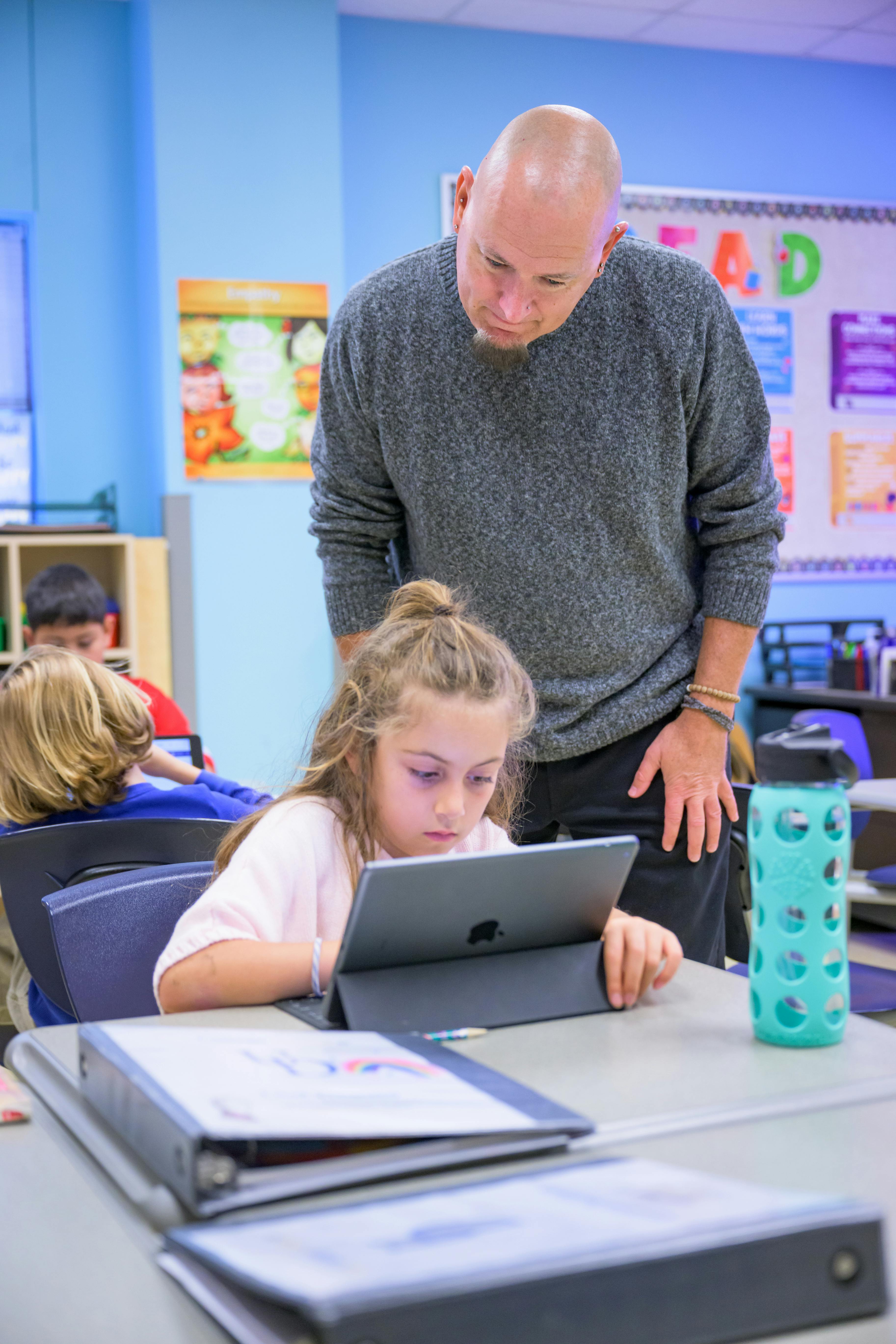 Adult standing over child using a tablet in a classroom