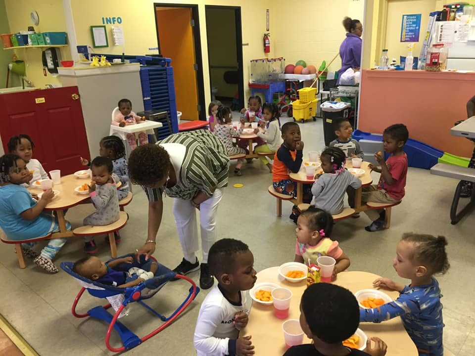 Adult interacting with children seated at tables in a classroom