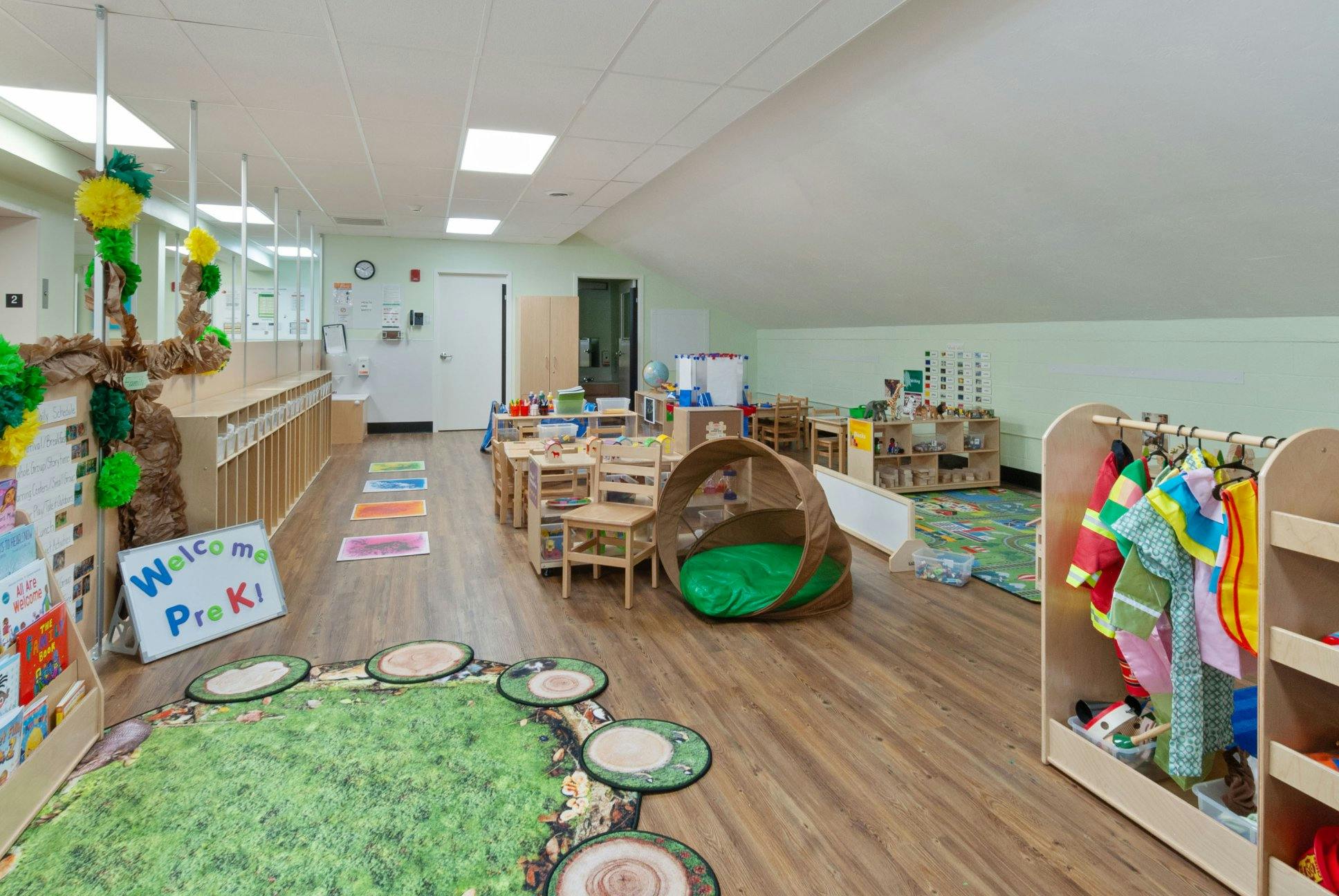 An empty classroom with small tables and chairs, a play tent, and a dress-up rack
