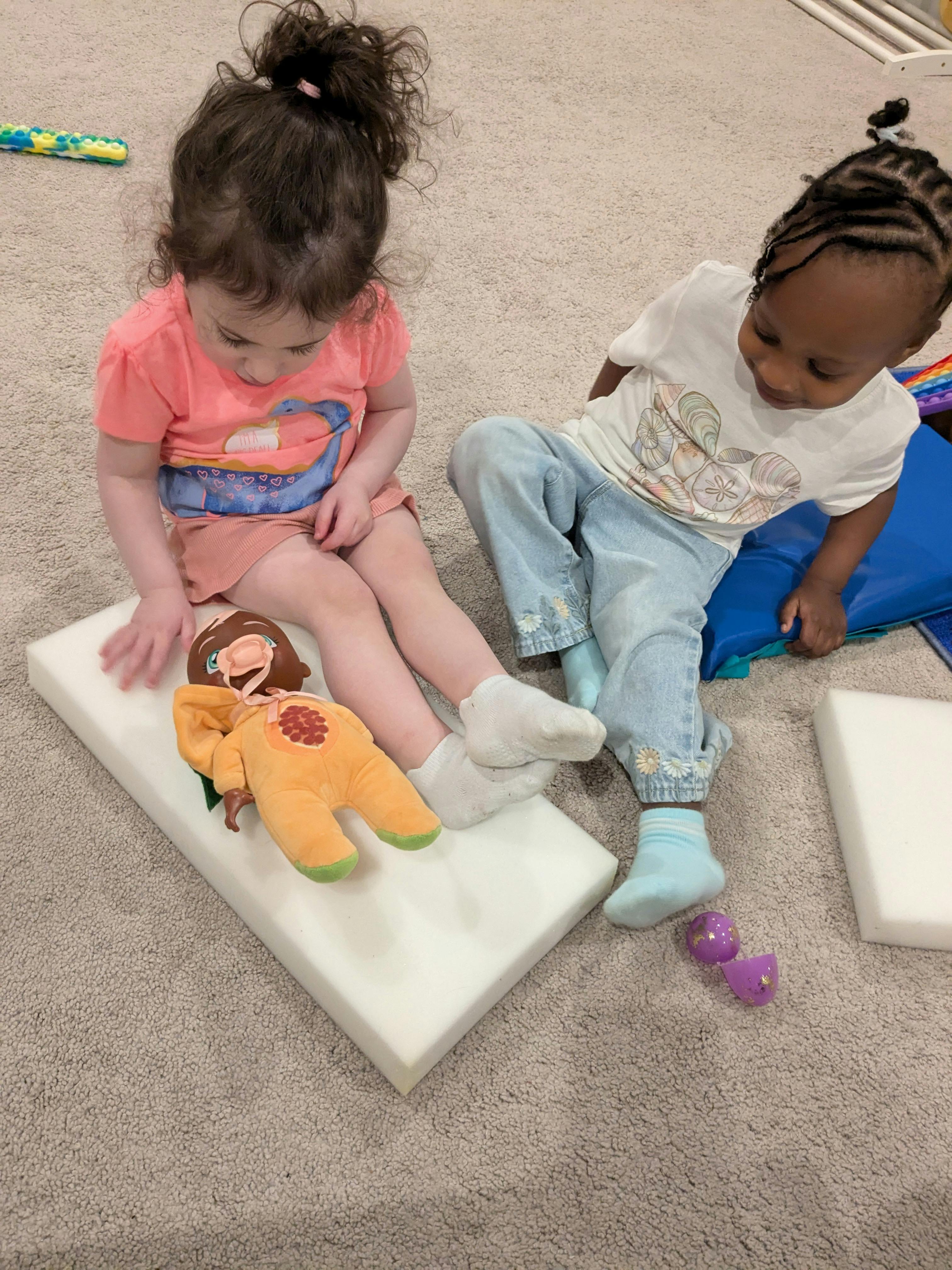 Two children sitting on the floor with a doll and foam blocks