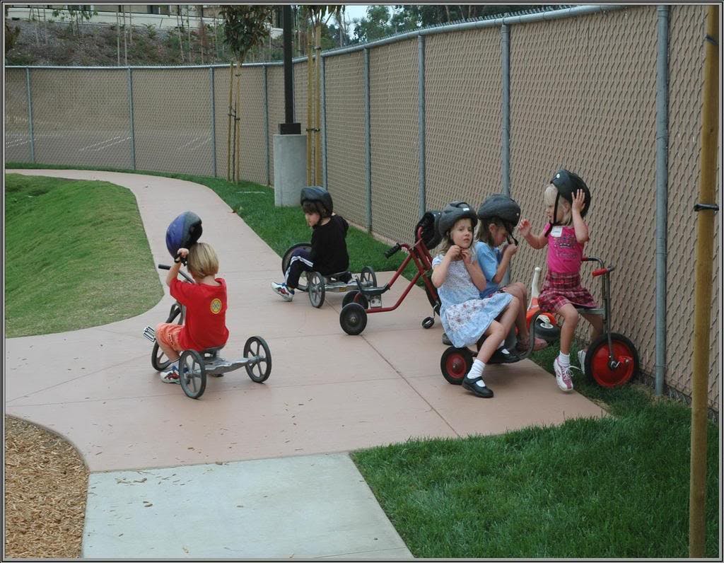 Children wearing helmets riding and sitting on tricycles on a paved path
