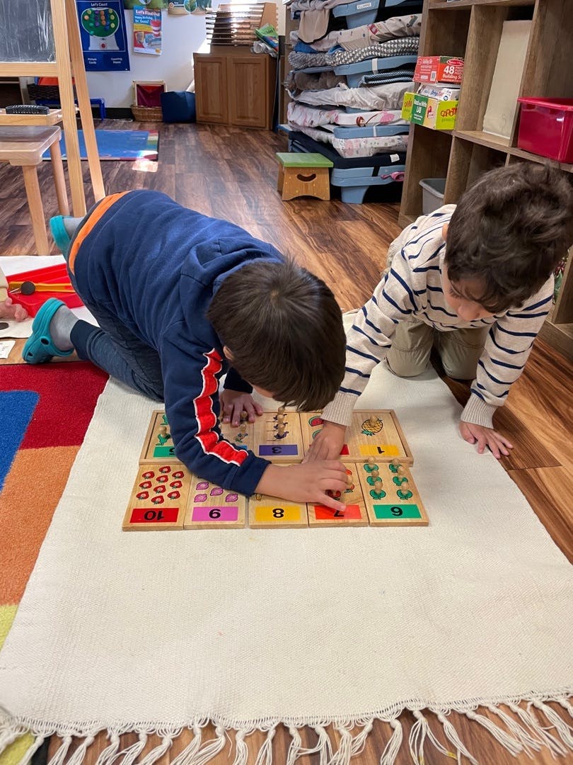 Two children kneeling on a rug, playing with a wooden number puzzle