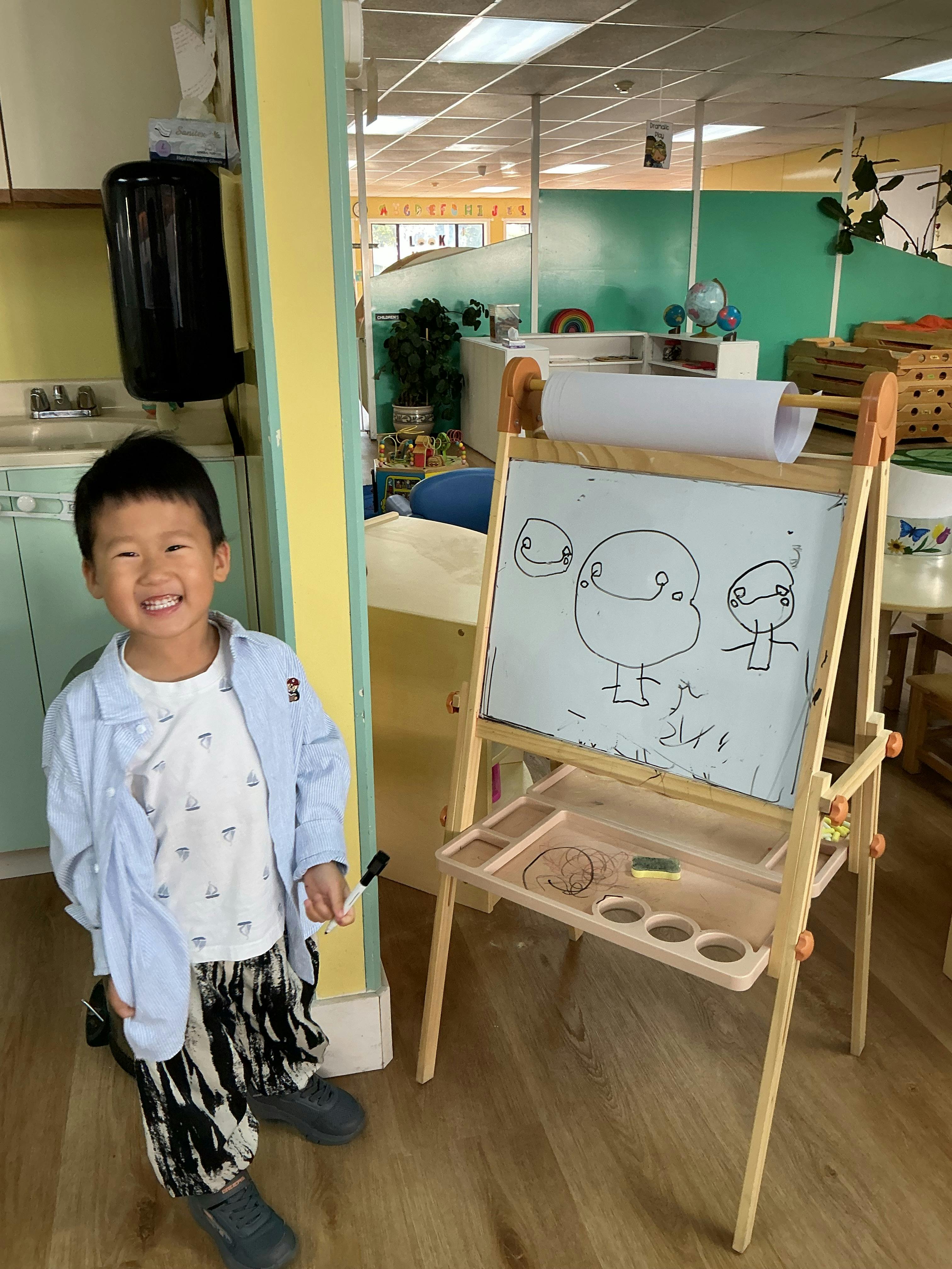 Child standing next to an easel with drawings on the whiteboard
