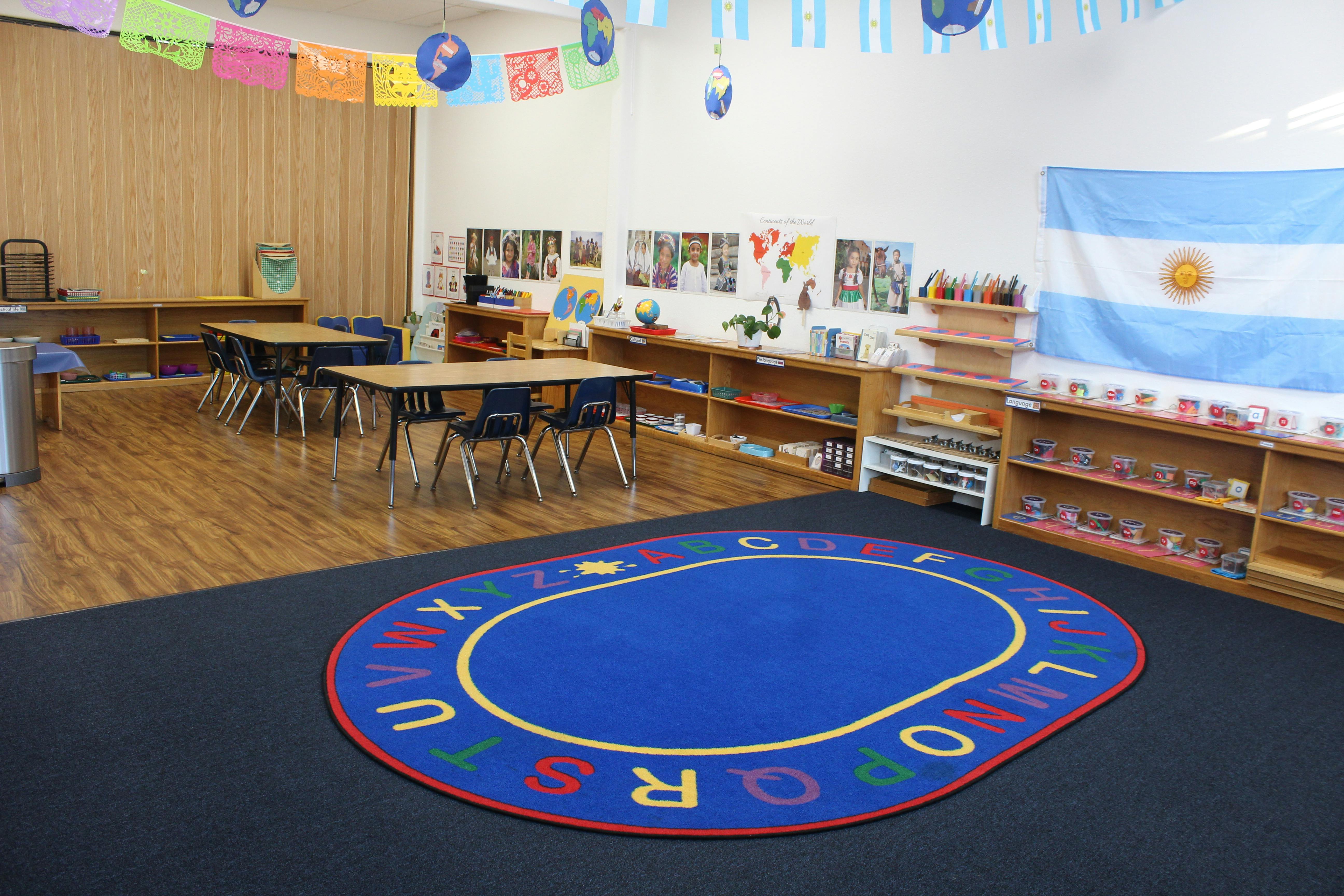 Classroom with small tables and chairs, shelves, and an alphabet rug