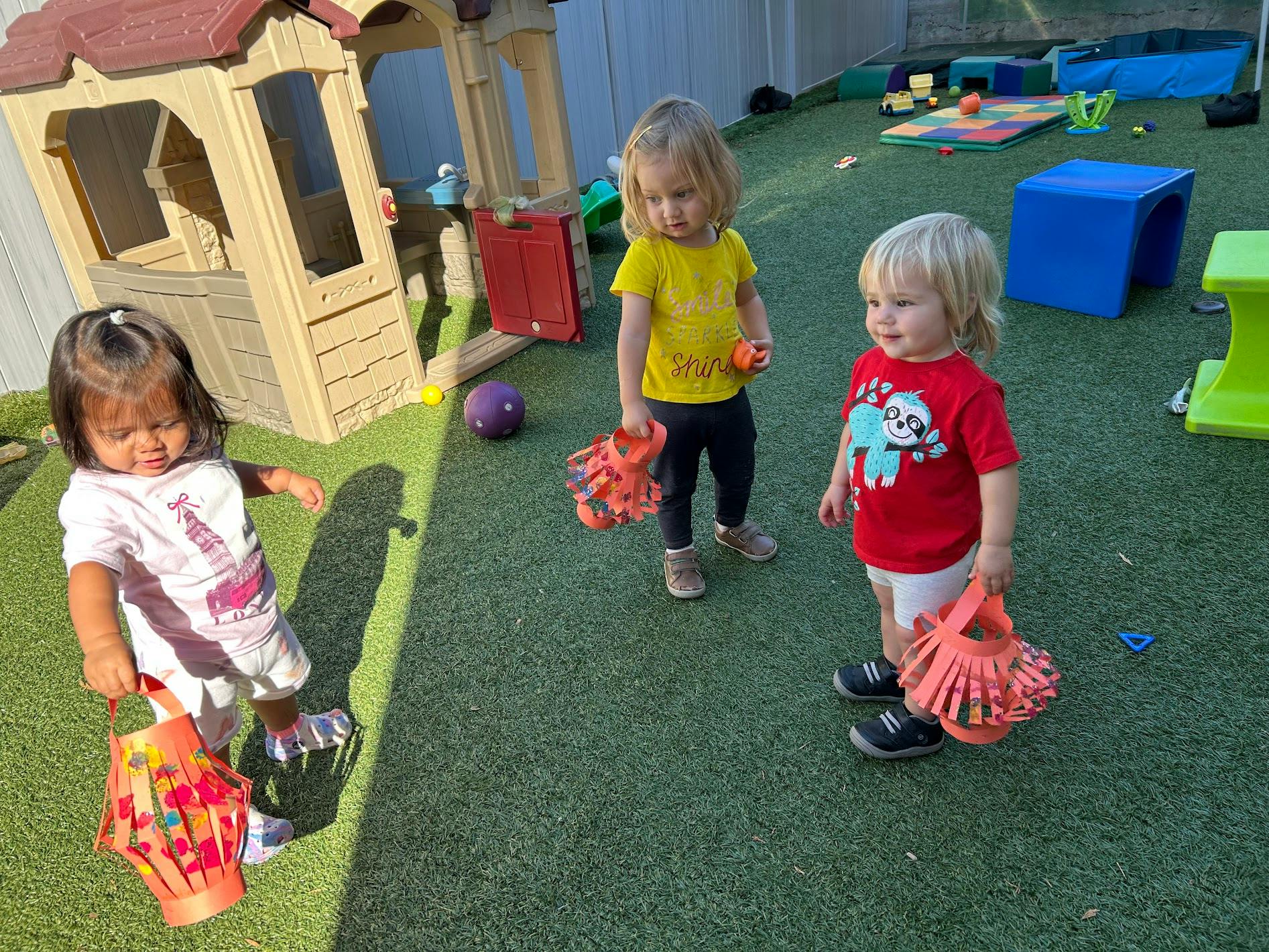 Three children holding handmade orange paper lanterns in a play area