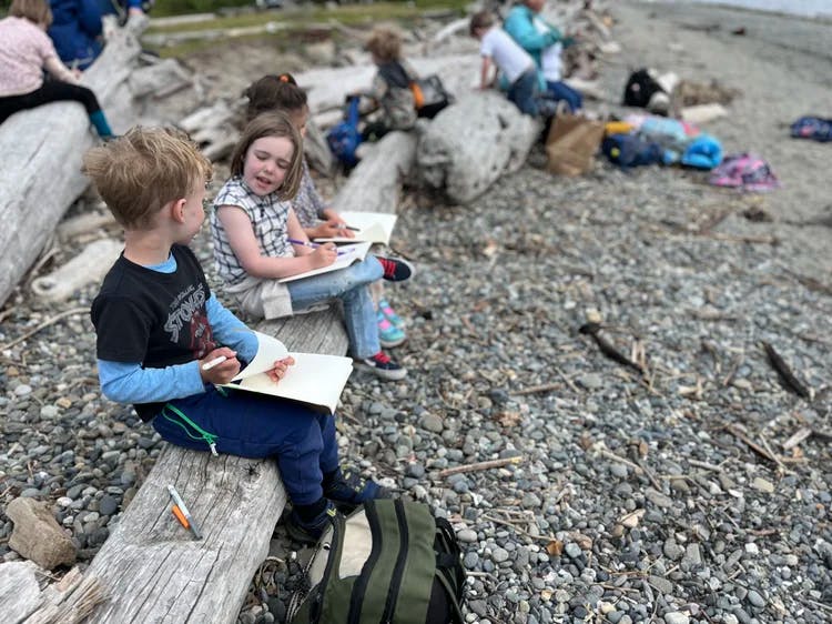 Children sitting on logs on a rocky beach writing in notebooks