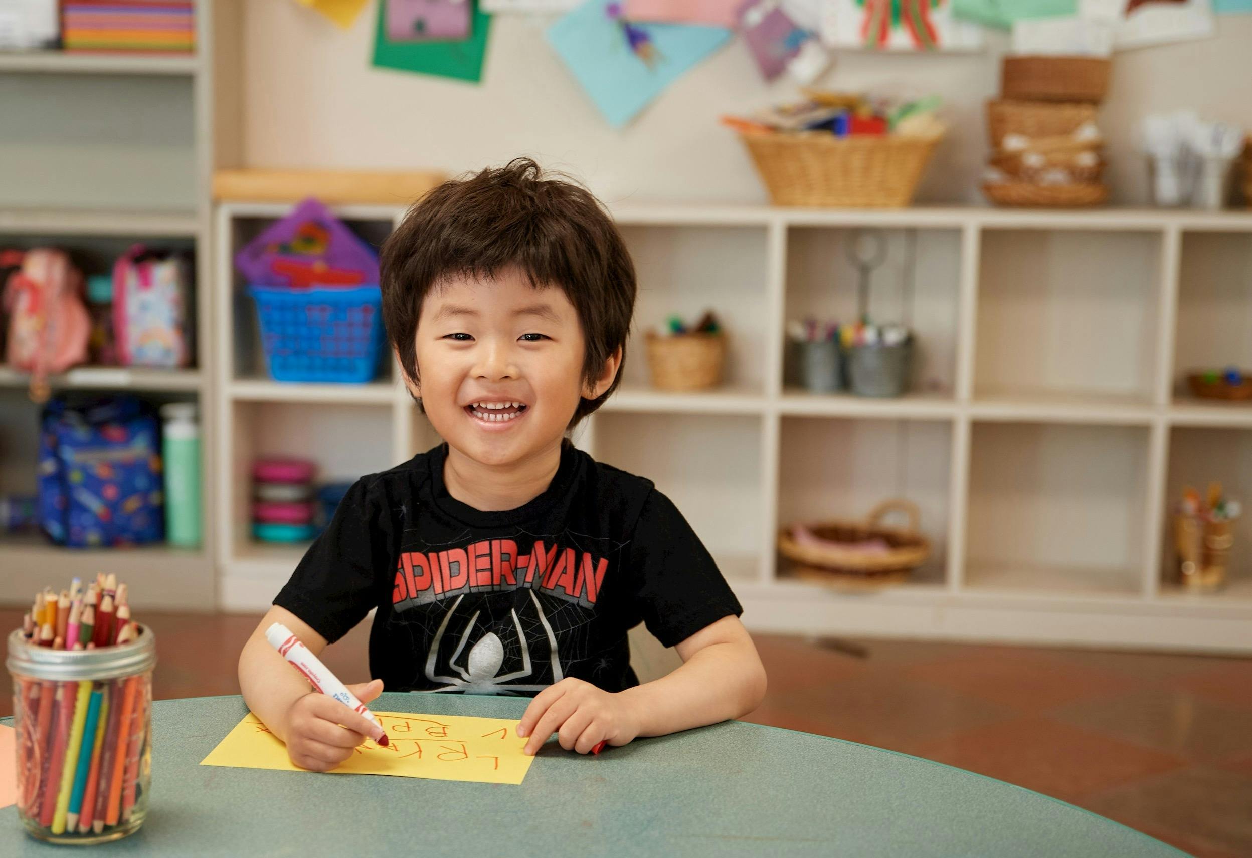 Child smiling while writing on yellow paper with a red marker in a classroom