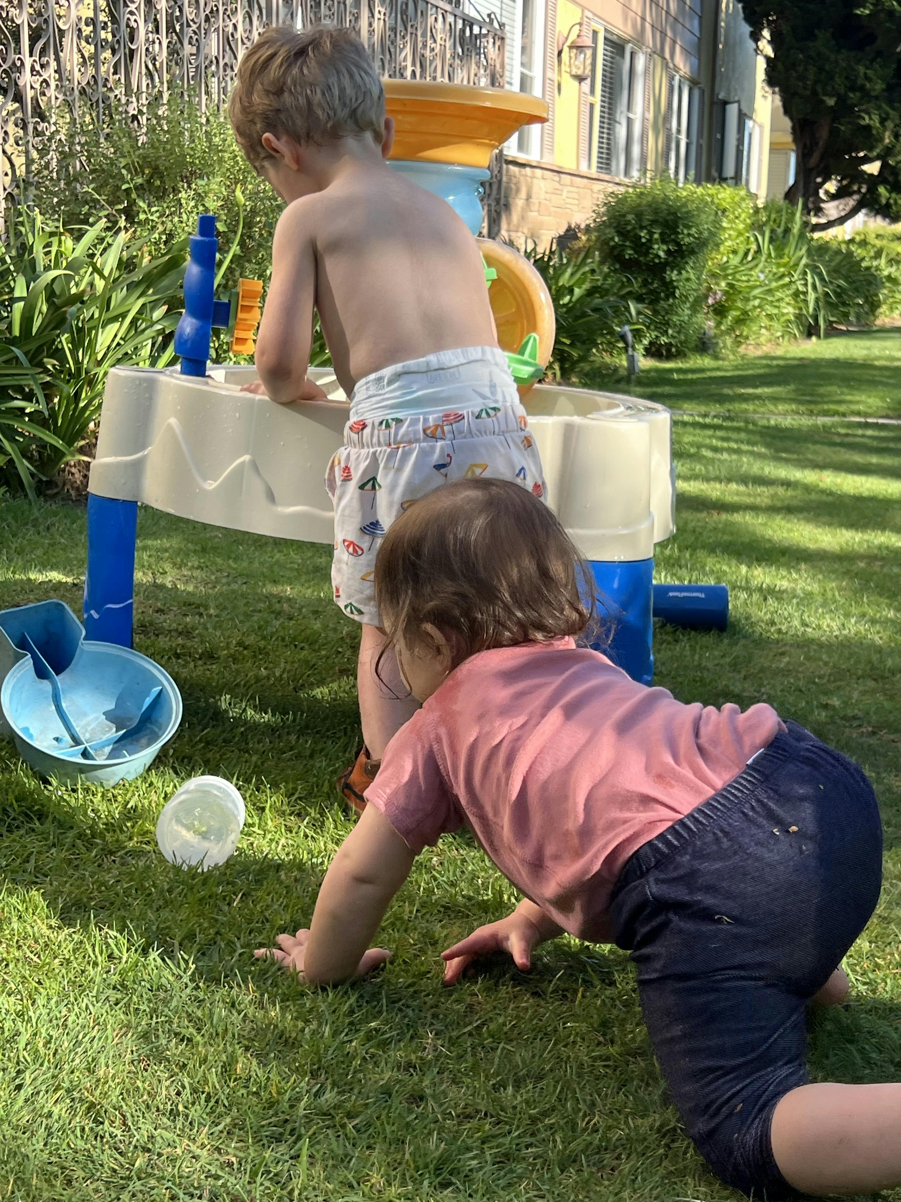 Child playing at a water table with another child crawling on the grass nearby grass
