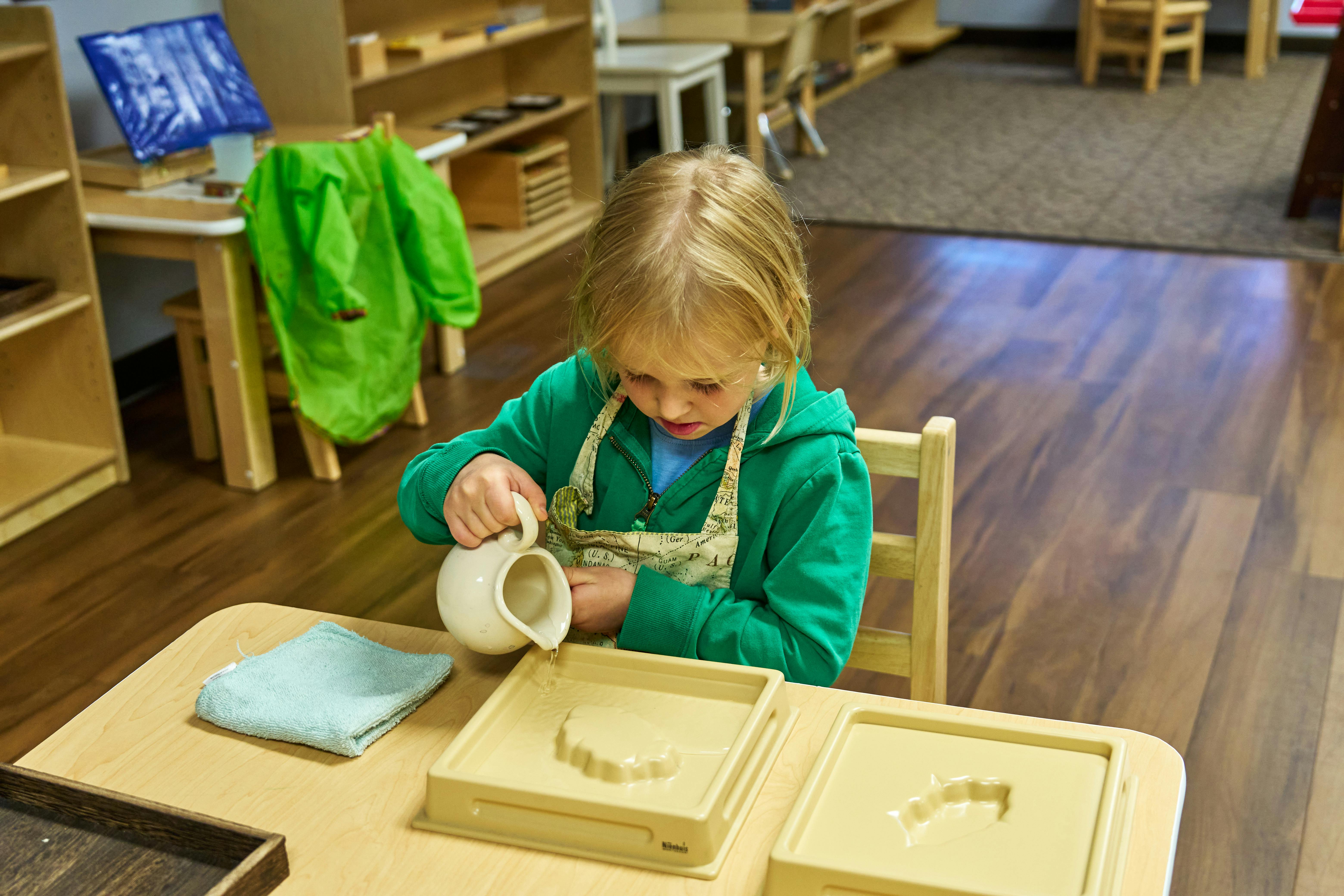Child pouring from a small pitcher into a tray at a table
