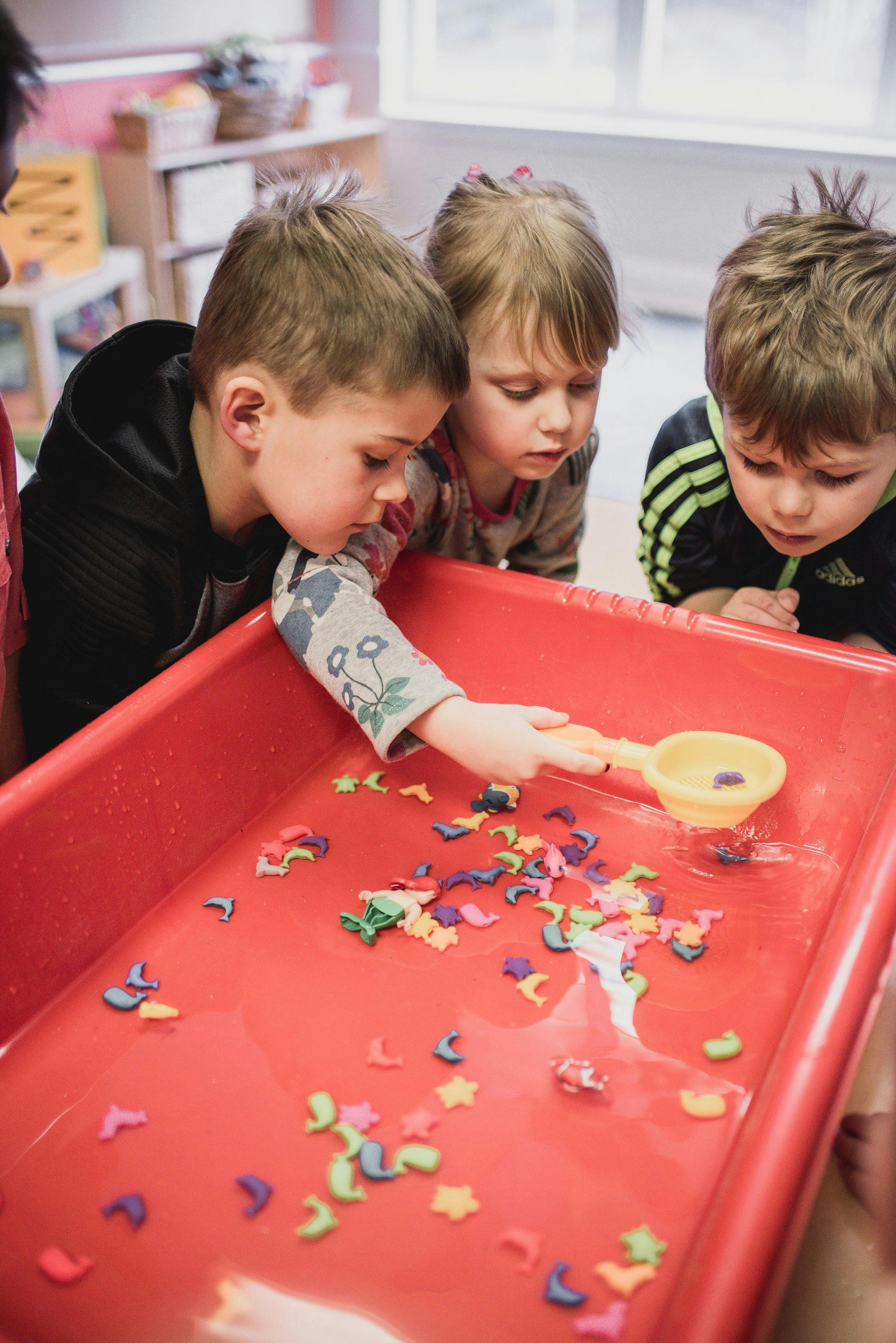 Three children playing with colorful shapes in a red bin