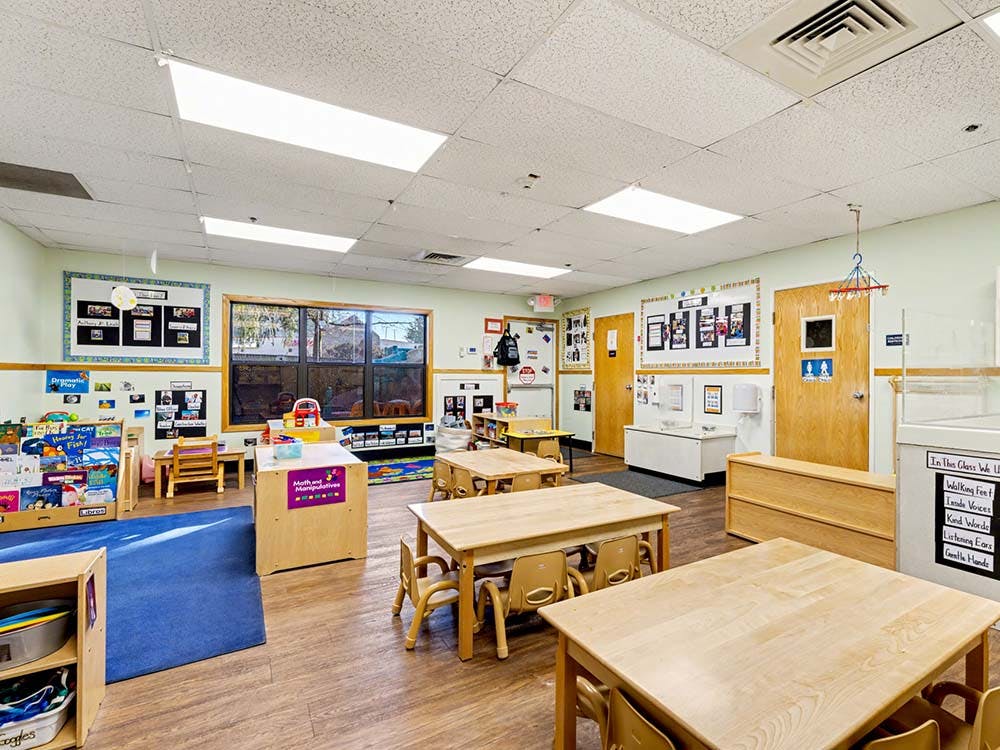 Empty classroom with small tables and chairs, shelves with books and toys