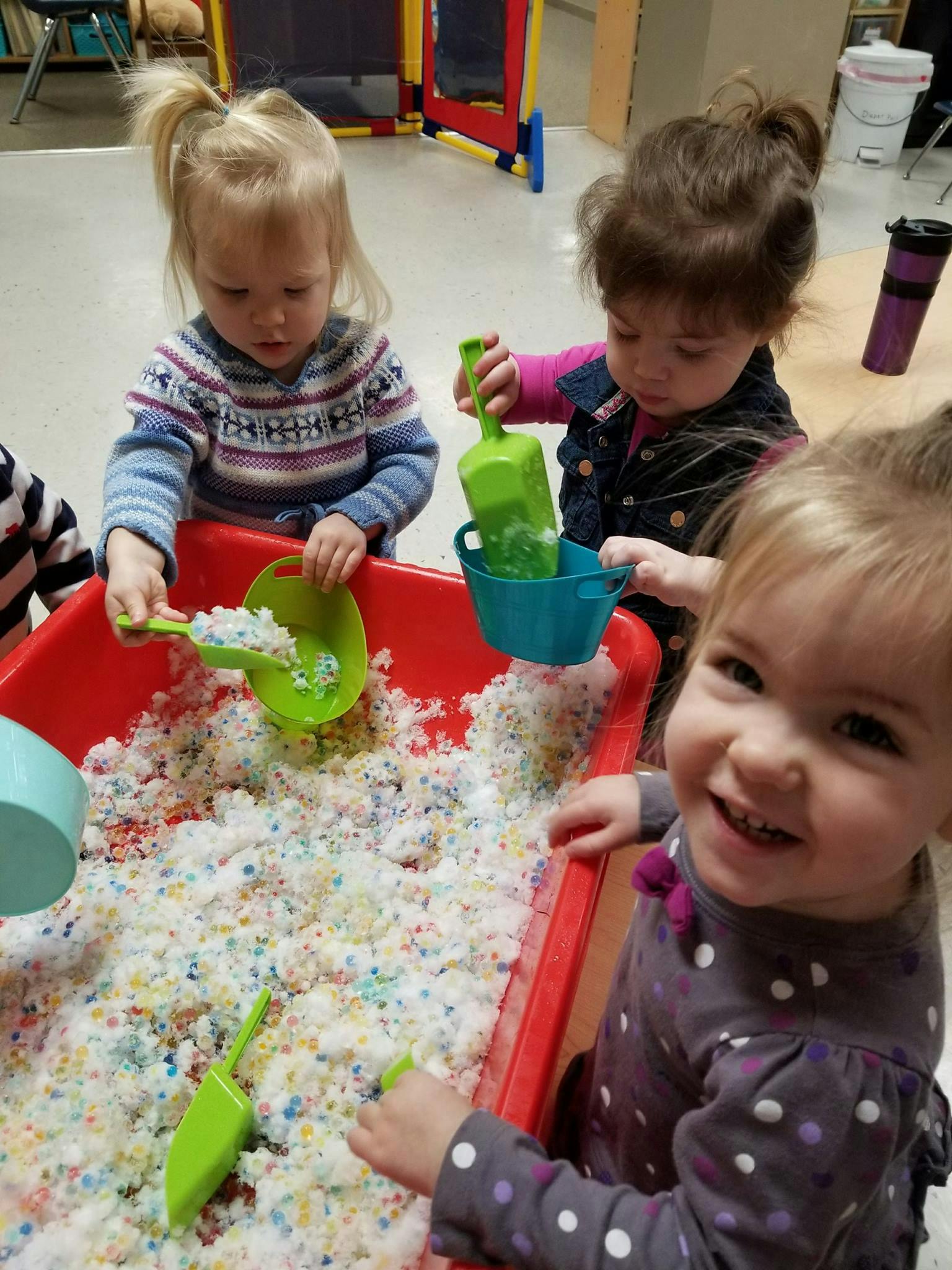 Three children playing with scoops and cups in a sensory bin with white and colorful beads