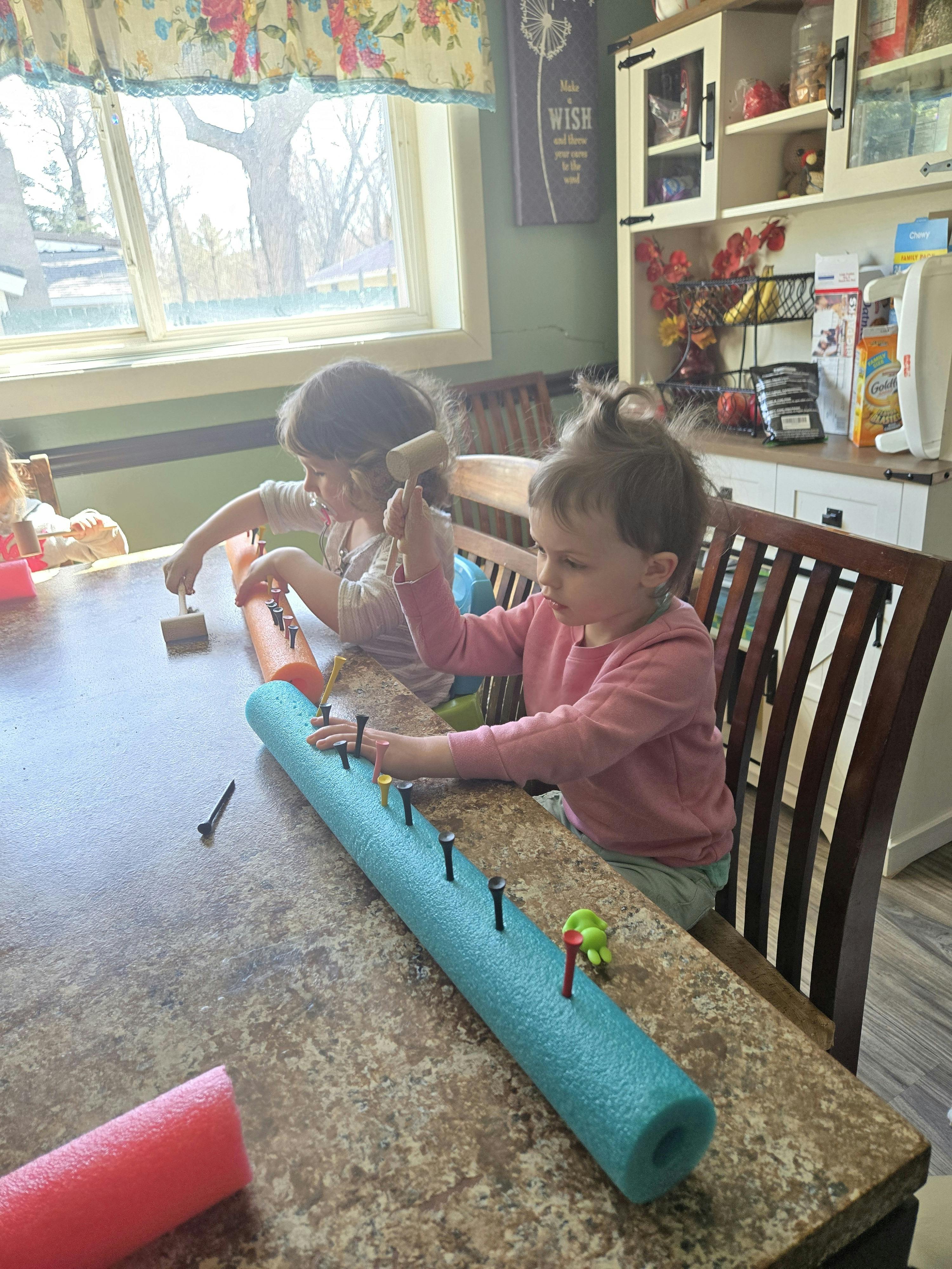 Two children hammering nails into pool noodles on a table