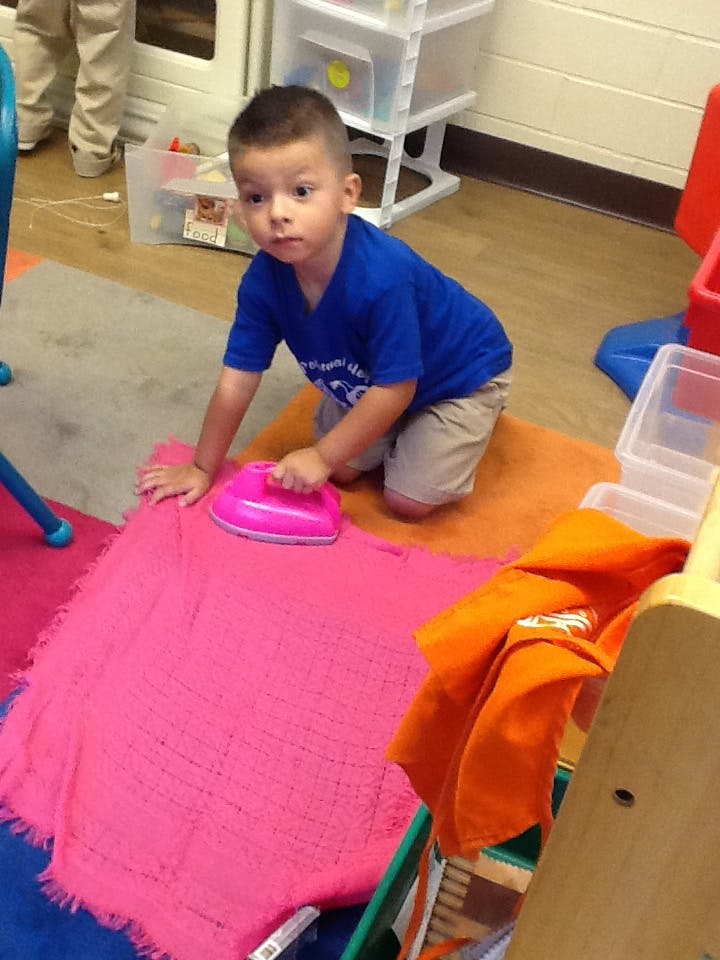 Child kneeling on floor, playing with a toy iron on a pink blanket