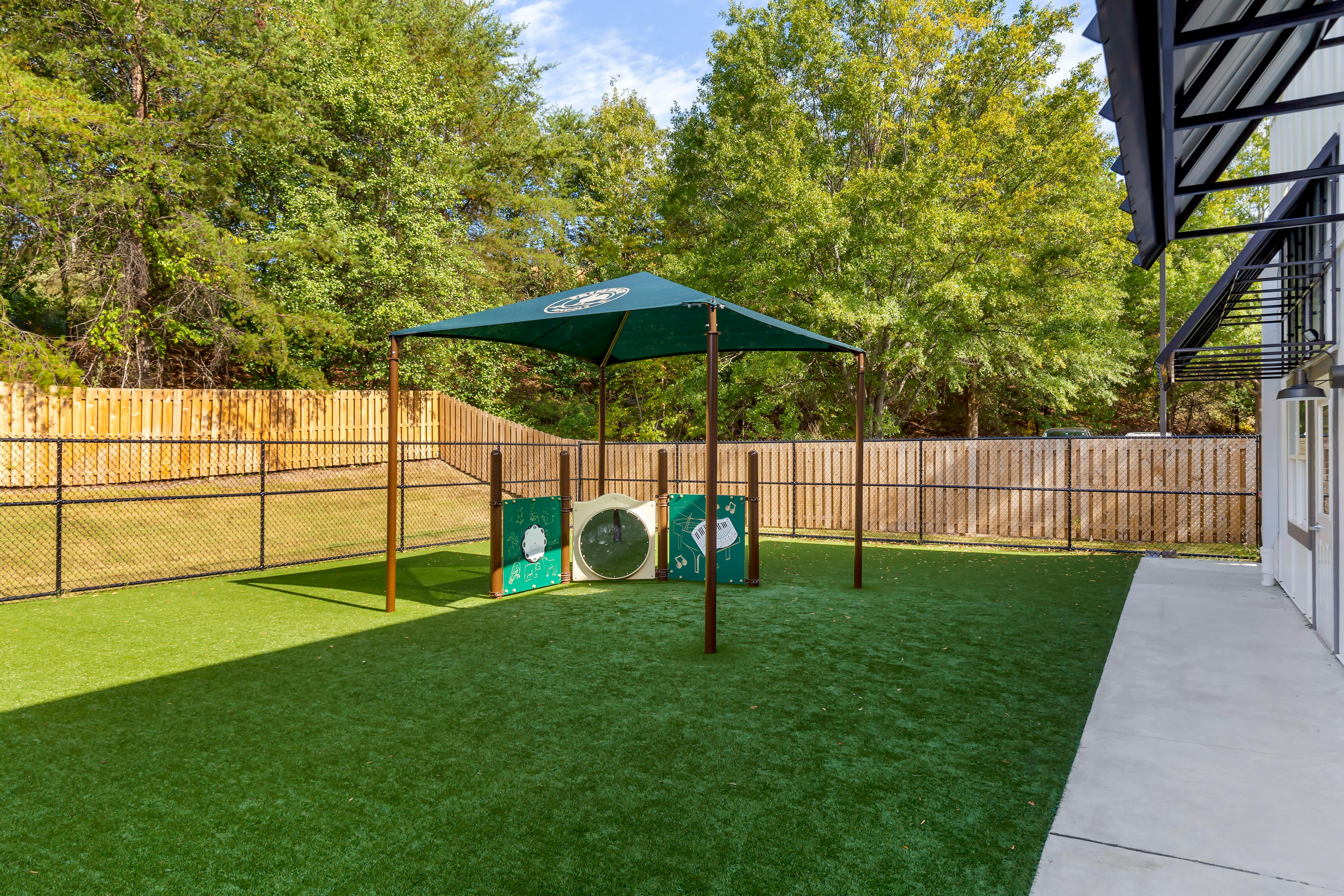 Artificial grass area with a green canopy and activity panels, fenced, next to a building