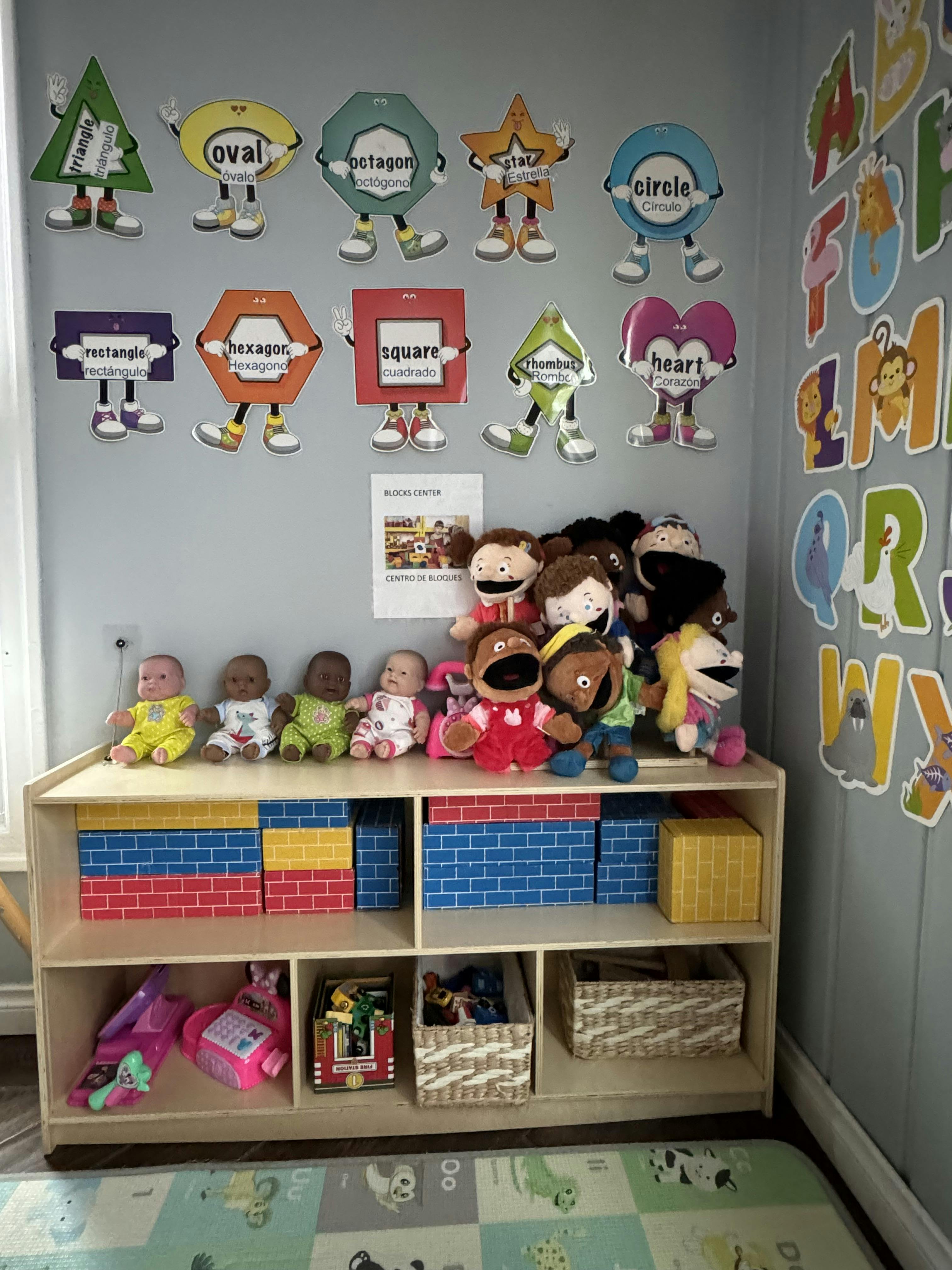 Classroom shelf with dolls, puppets, and boxes under shape cutouts and alphabet letters