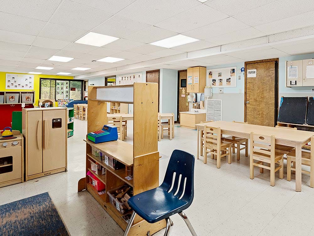 Empty classroom with small wooden tables and chairs, play kitchen, and register