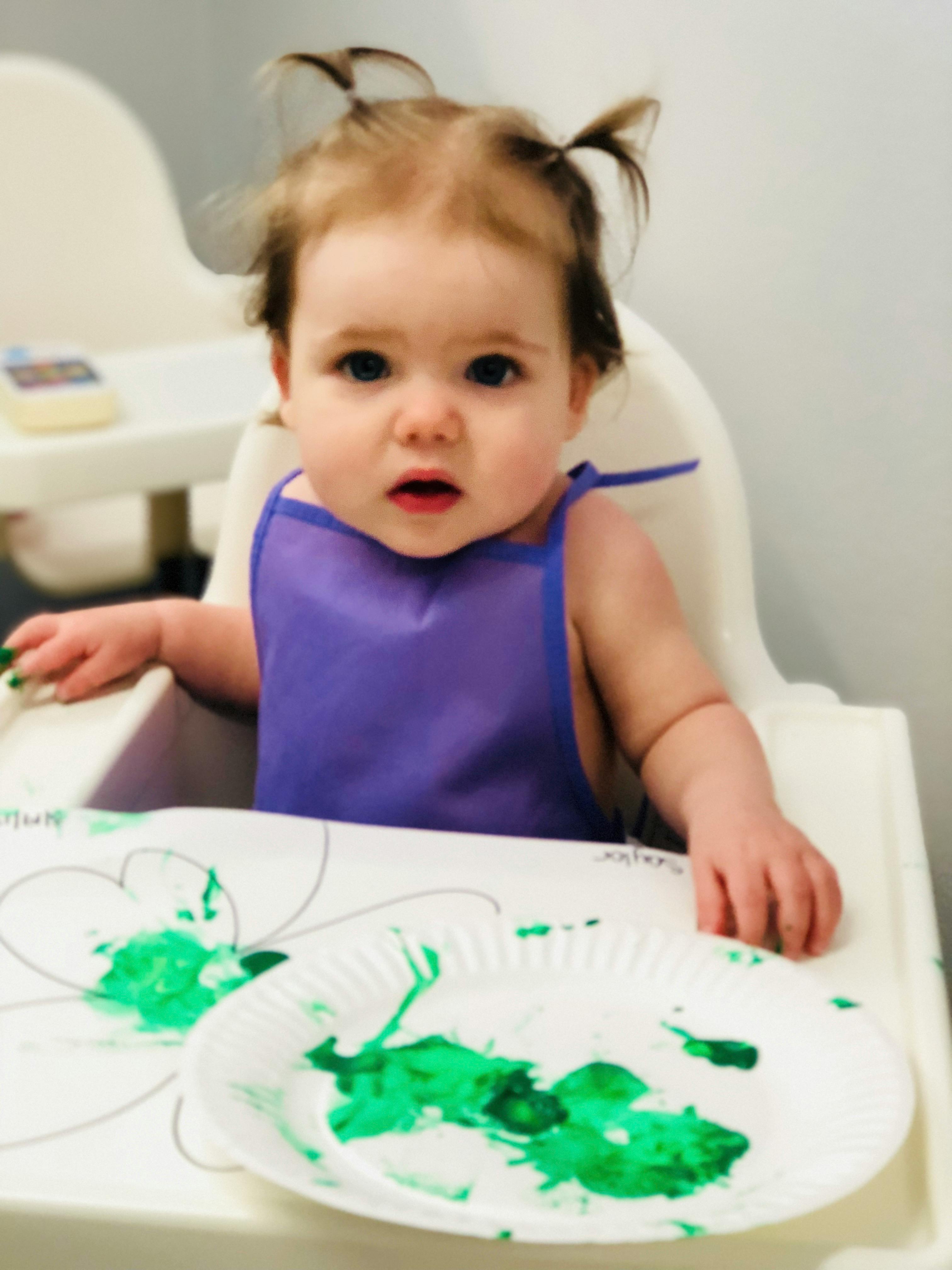 Child in a high chair with a purple bib and green food on a paper plate