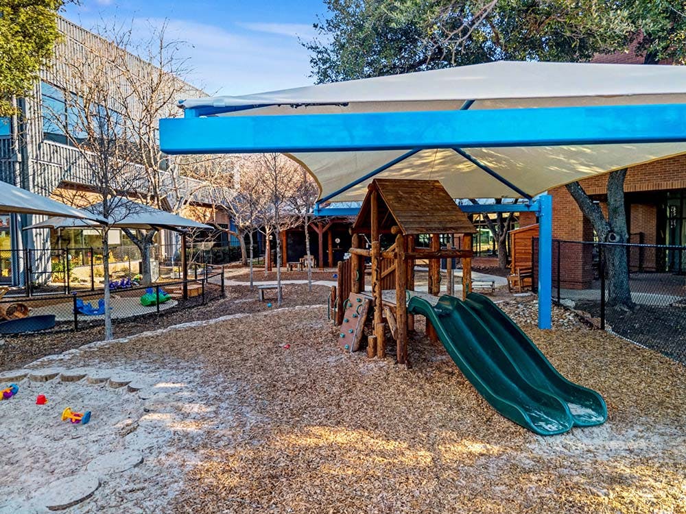 Outdoor children's playground with a wooden climbing structure, slide, sand area, and shade cover