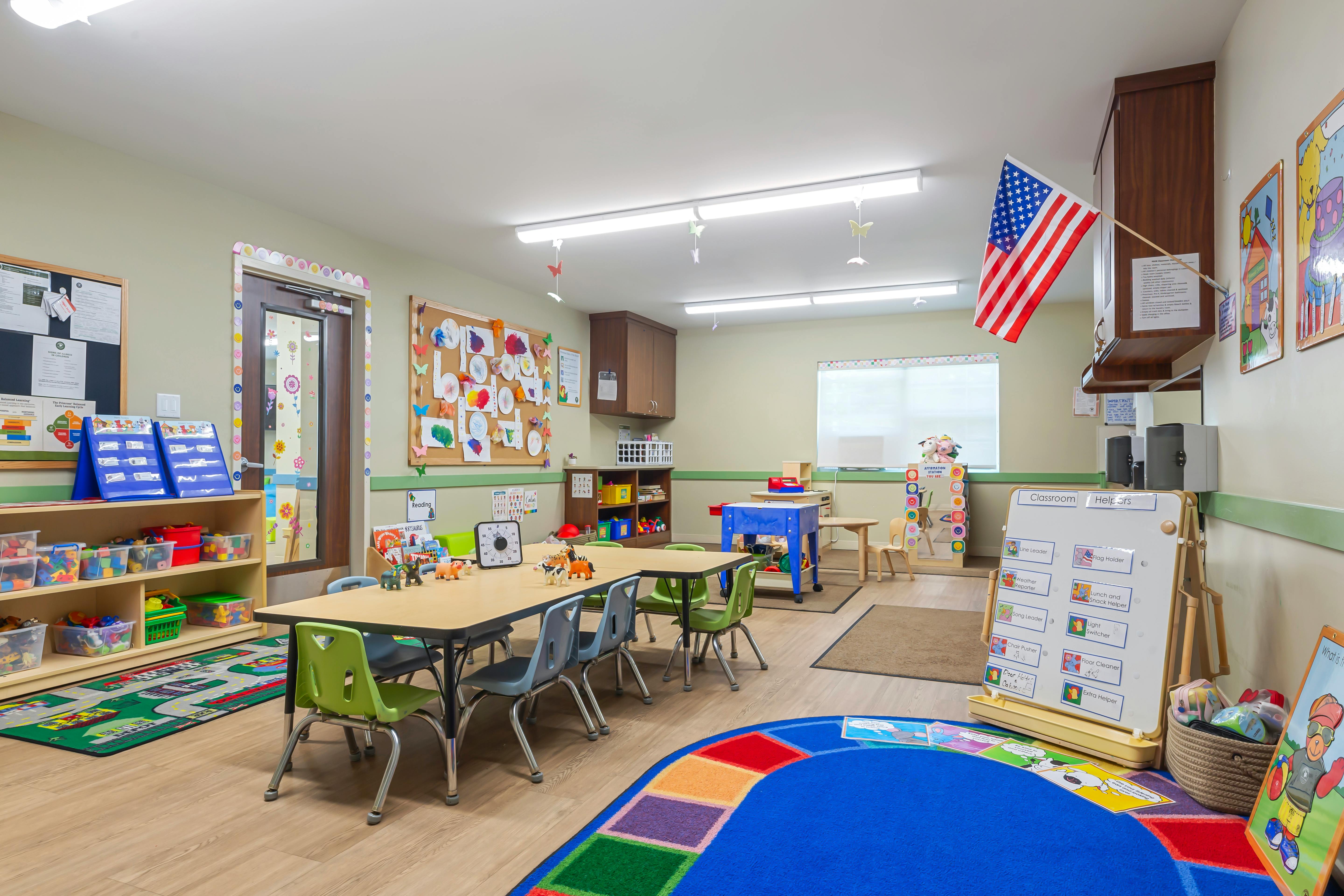 Colorful classroom with small tables and chairs, a play rug, and classroom helper chart