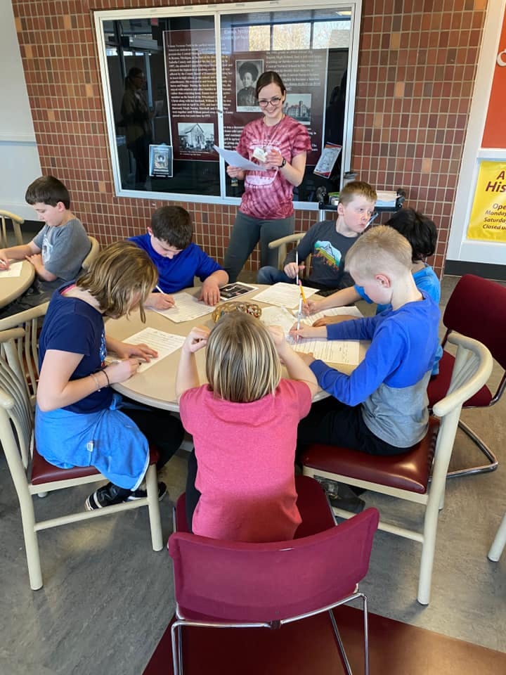 Adult person standing and children sitting at tables writing