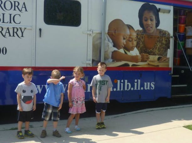 Four children standing in front of a bookmobile with a mural of a person reading to children