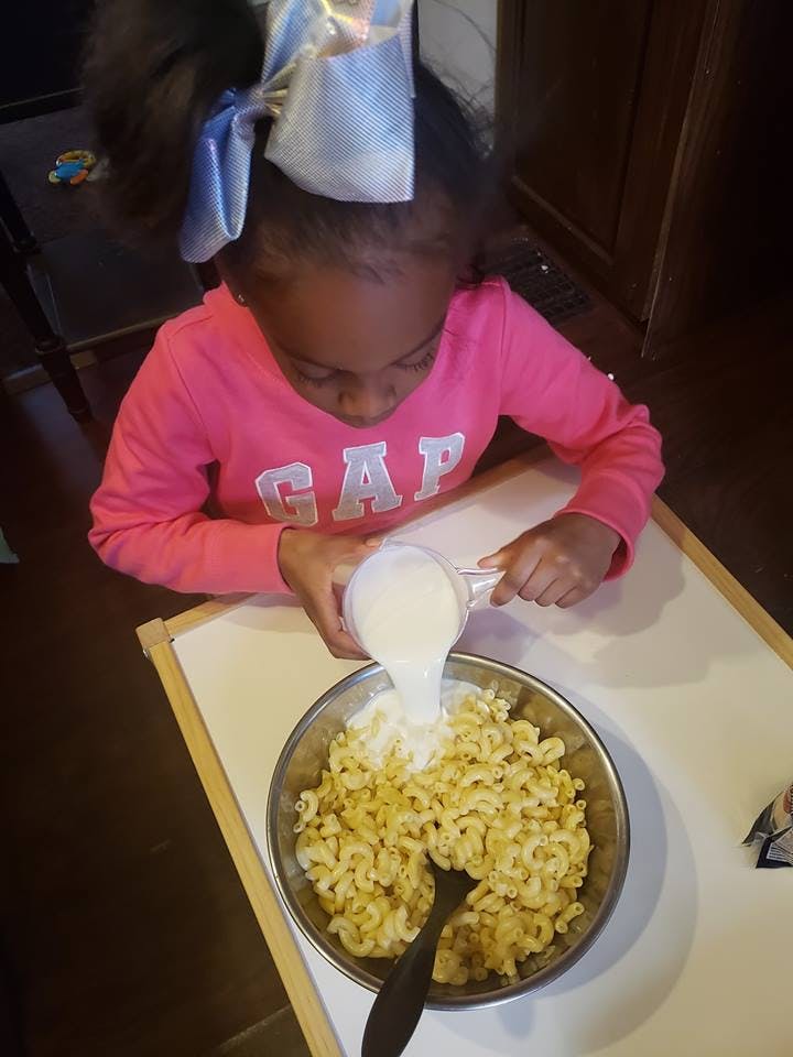 Child pouring liquid into a bowl of macaroni
