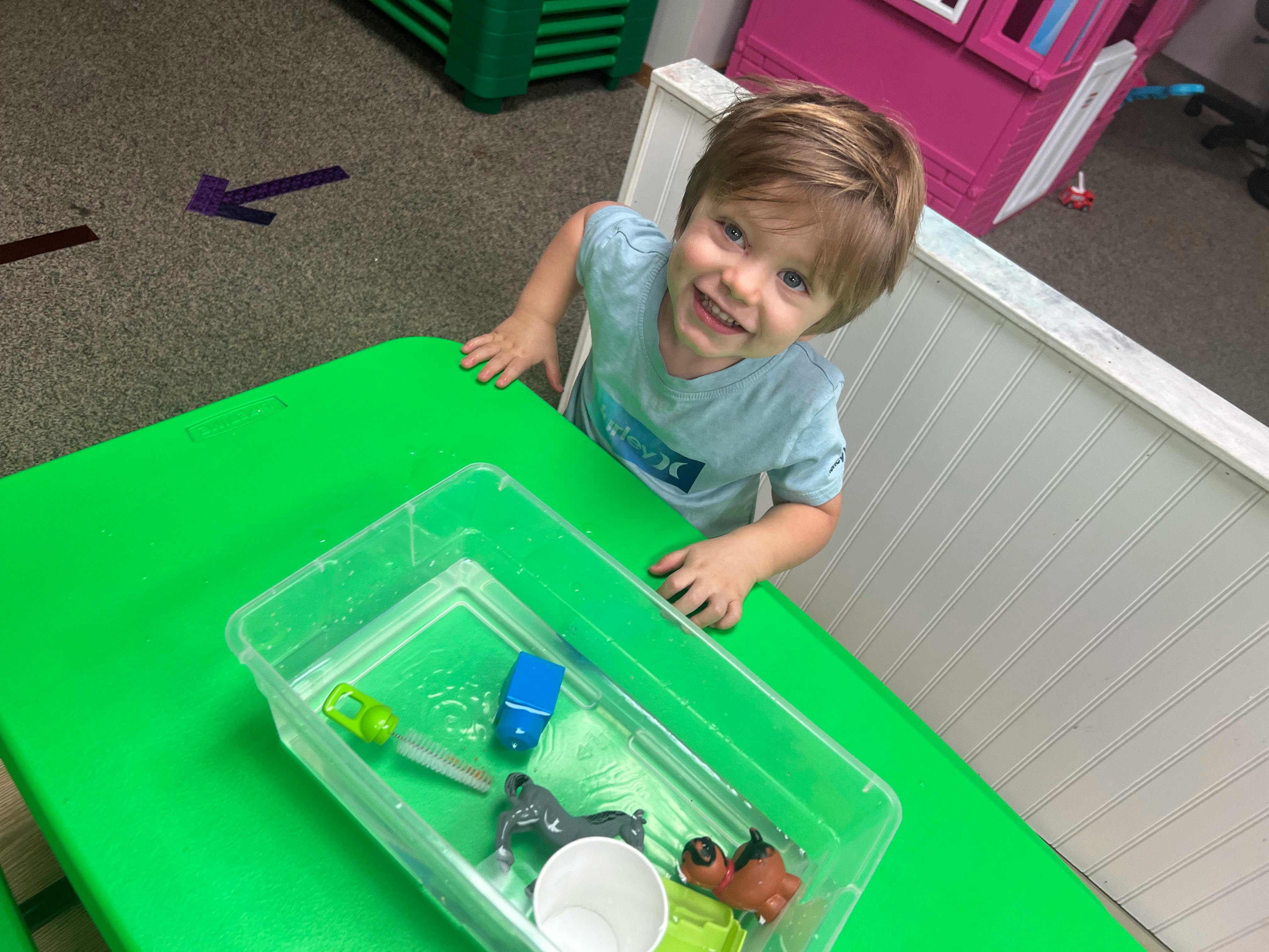 Child looking up from a table with a clear bin of toys