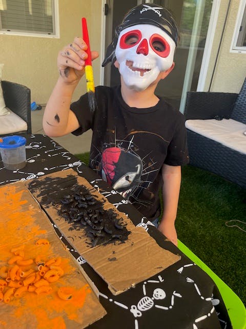 Child in a pirate skull mask holding a paintbrush over painted cardboard