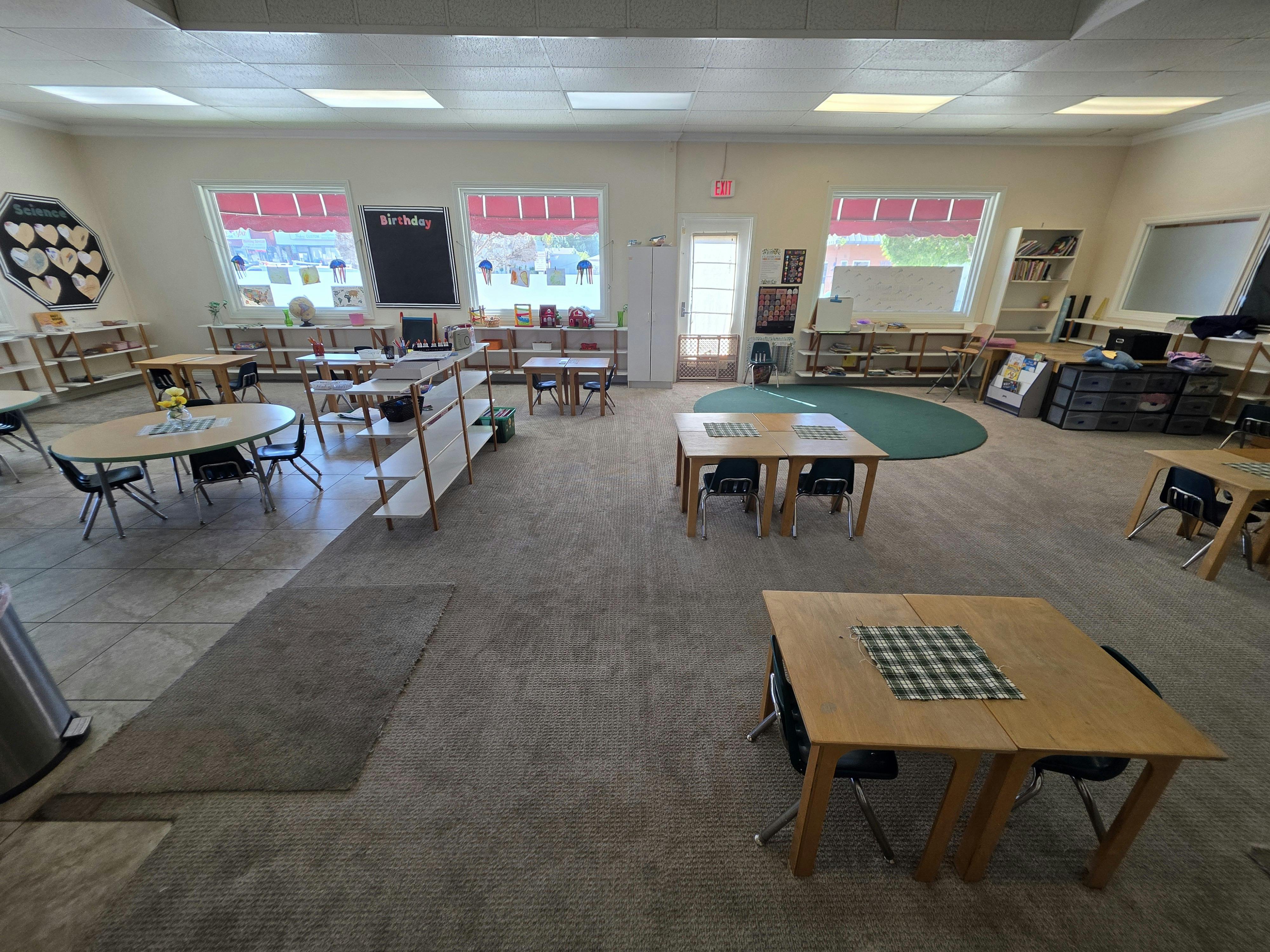 An empty classroom with small tables and chairs, shelves, and windows