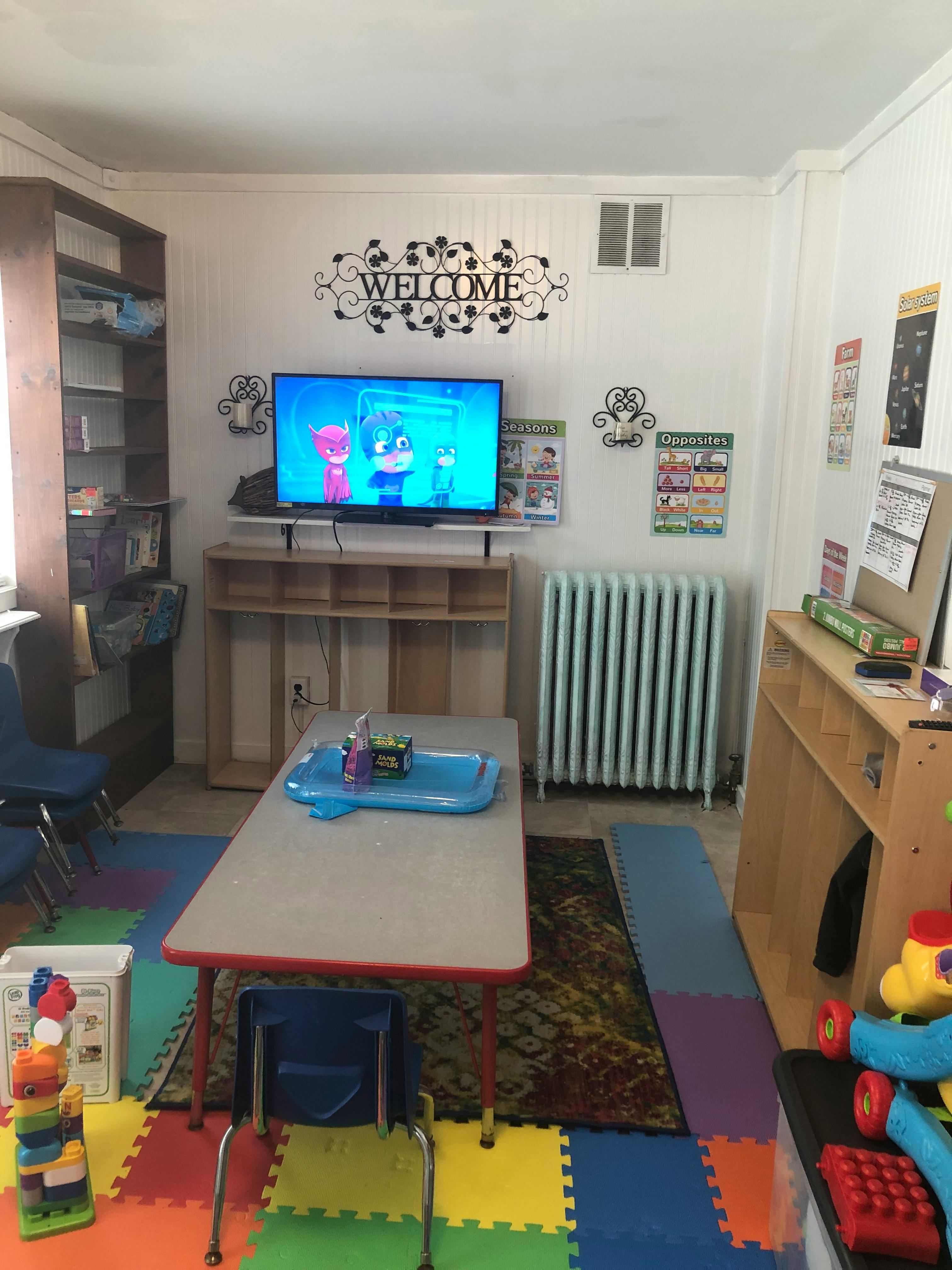 Classroom with a TV displaying cartoons, a table, and colorful floor mats