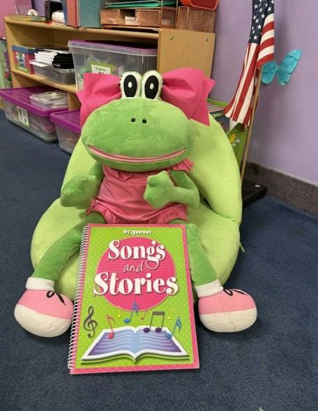 Plush frog with a bow holding a Songs and Stories book in a classroom