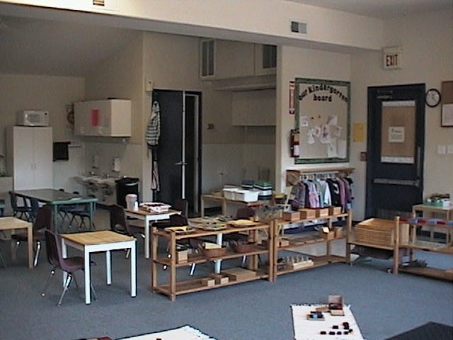 Classroom with small tables and chairs, wooden shelves with learning materials
