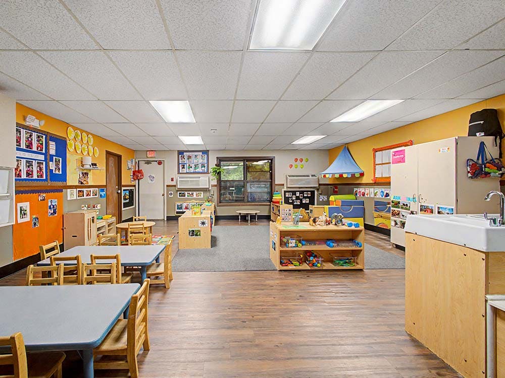Empty classroom with small tables and chairs, shelves with toys, and a sink