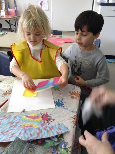 Two children at a table, one holding colorful paper over a white sheet