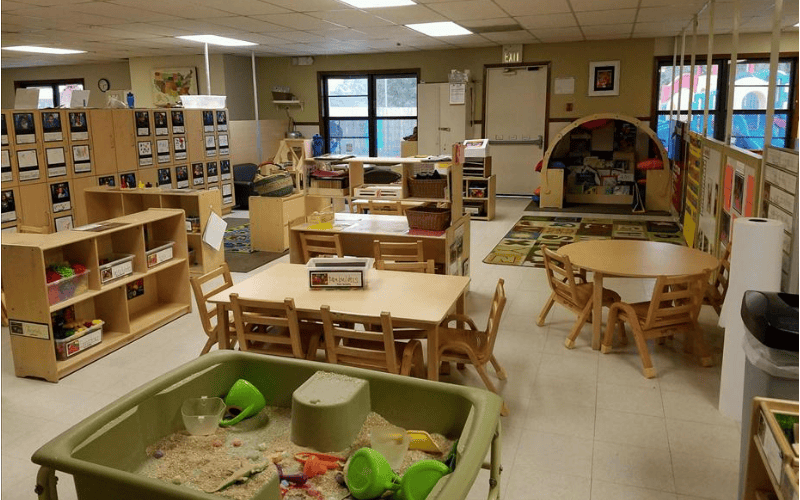 An empty classroom with small tables and chairs, shelves, and a sand table