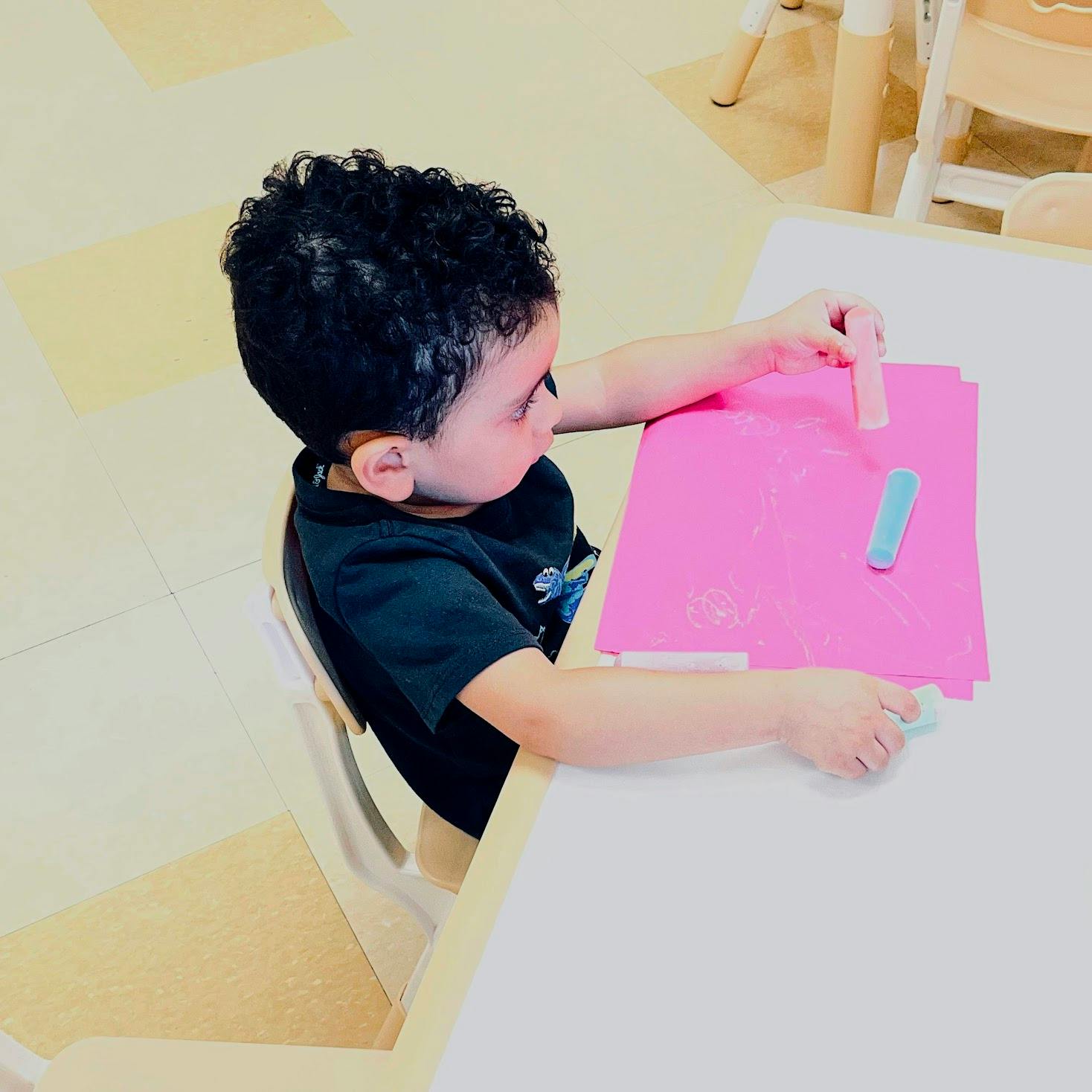 Child drawing with chalk on pink paper at a table