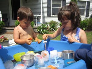Two children playing with ice and water in a blue bin