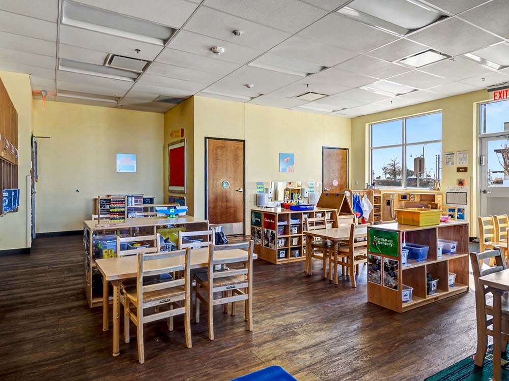 Empty classroom with small tables, chairs, and shelves with toys and learning materials