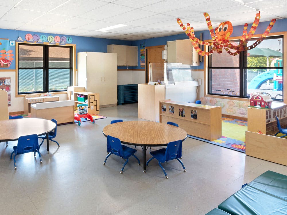 Empty toddler classroom with small tables and chairs, play kitchen, and paper octopus