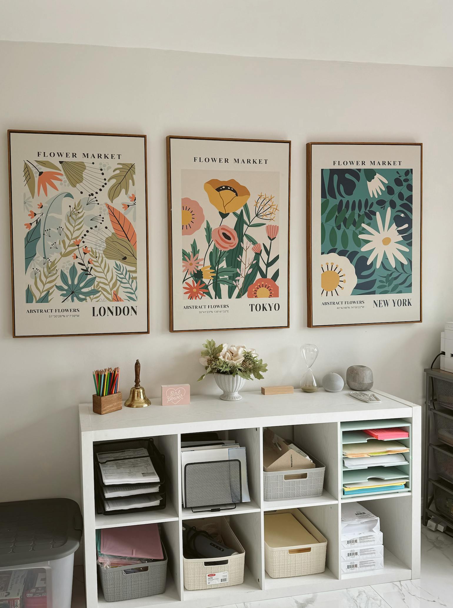 Three framed flower market posters above a white storage shelf with office supplies