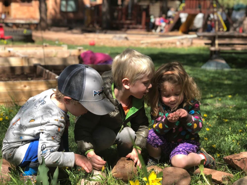 Three children crouching in grass, looking at something on the ground