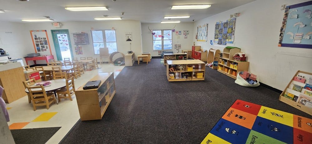 An empty classroom with small tables and chairs, shelves, and a colorful rug