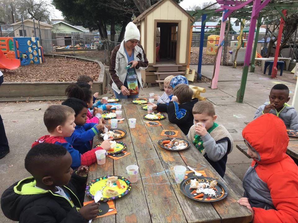 Person serving food to children at a long wooden table outdoors